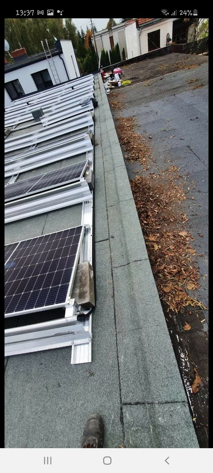 Panele słoneczne w trakcie montażu na płaskim dachu, zabezpieczone betonowymi blokami, widoczne liście i zabrudzenia na powierzchni dachu.