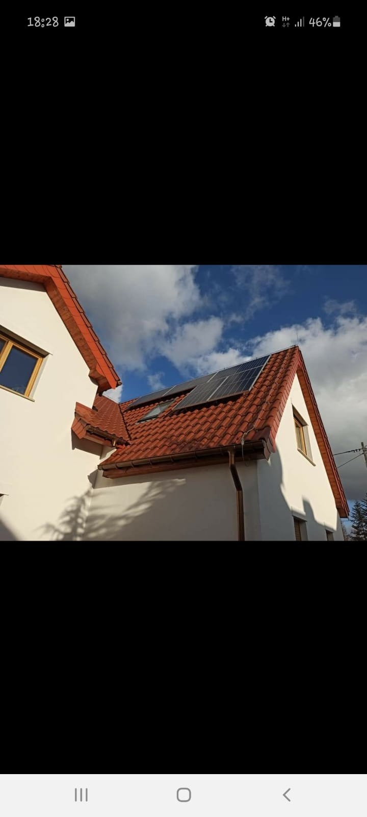 Panele słoneczne zamontowane na dachu domu z czerwoną dachówką, widoczne okna dachowe i system rynnowy, błękitne niebo z chmurami w tle.