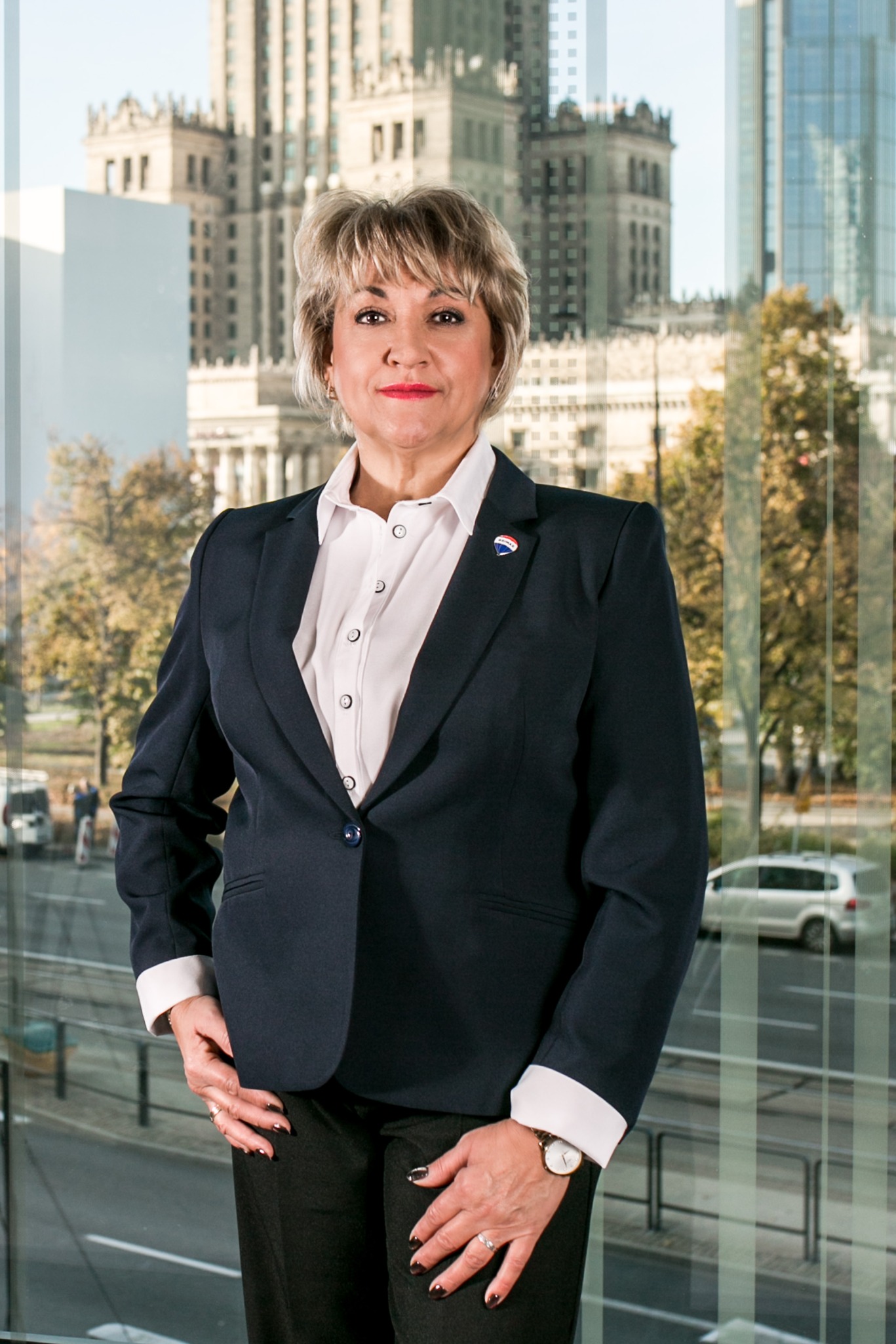 Profesjonalny portret kobiety w garniturze z przypinką agencji nieruchomości na tle biurowca i widoku na Warszawę.