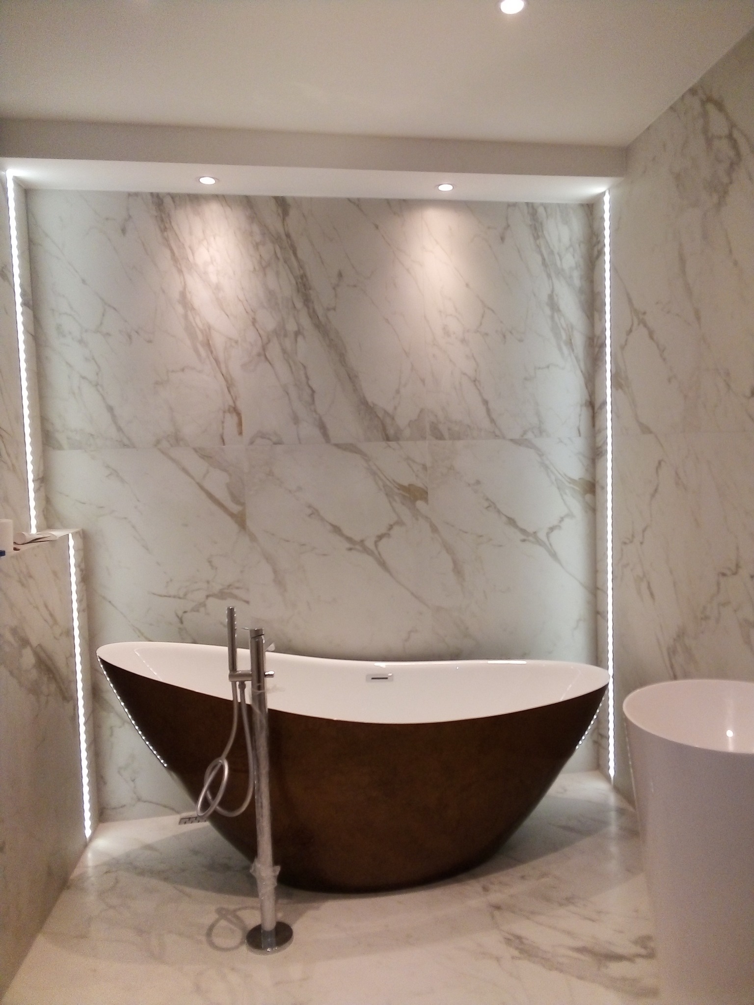 Nowoczesna łazienka z wolnostojącą, brązową wanną i marmurowymi płytkami ściennymi z subtelnym oświetleniem LED wzdłuż krawędzi.