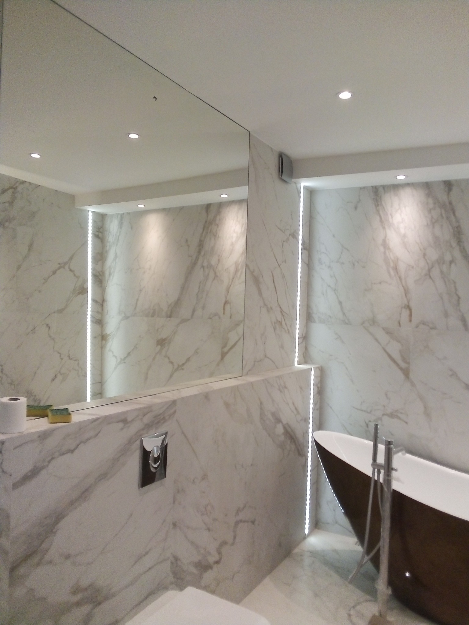 Nowoczesna łazienka z marmurowymi ścianami, podświetleniem LED wzdłuż krawędzi, wolnostojącą wanną w kolorze brązowym i dużym lustrem.