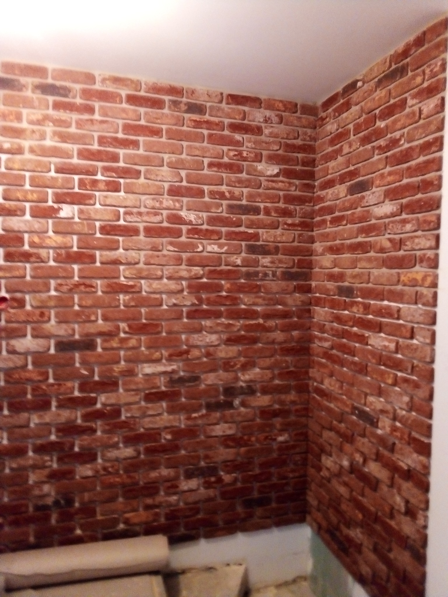 Fragment ściany wykończonej czerwoną cegłą dekoracyjną, widoczny róg pomieszczenia z niedokończoną podłogą i rolką materiału budowlanego w tle.