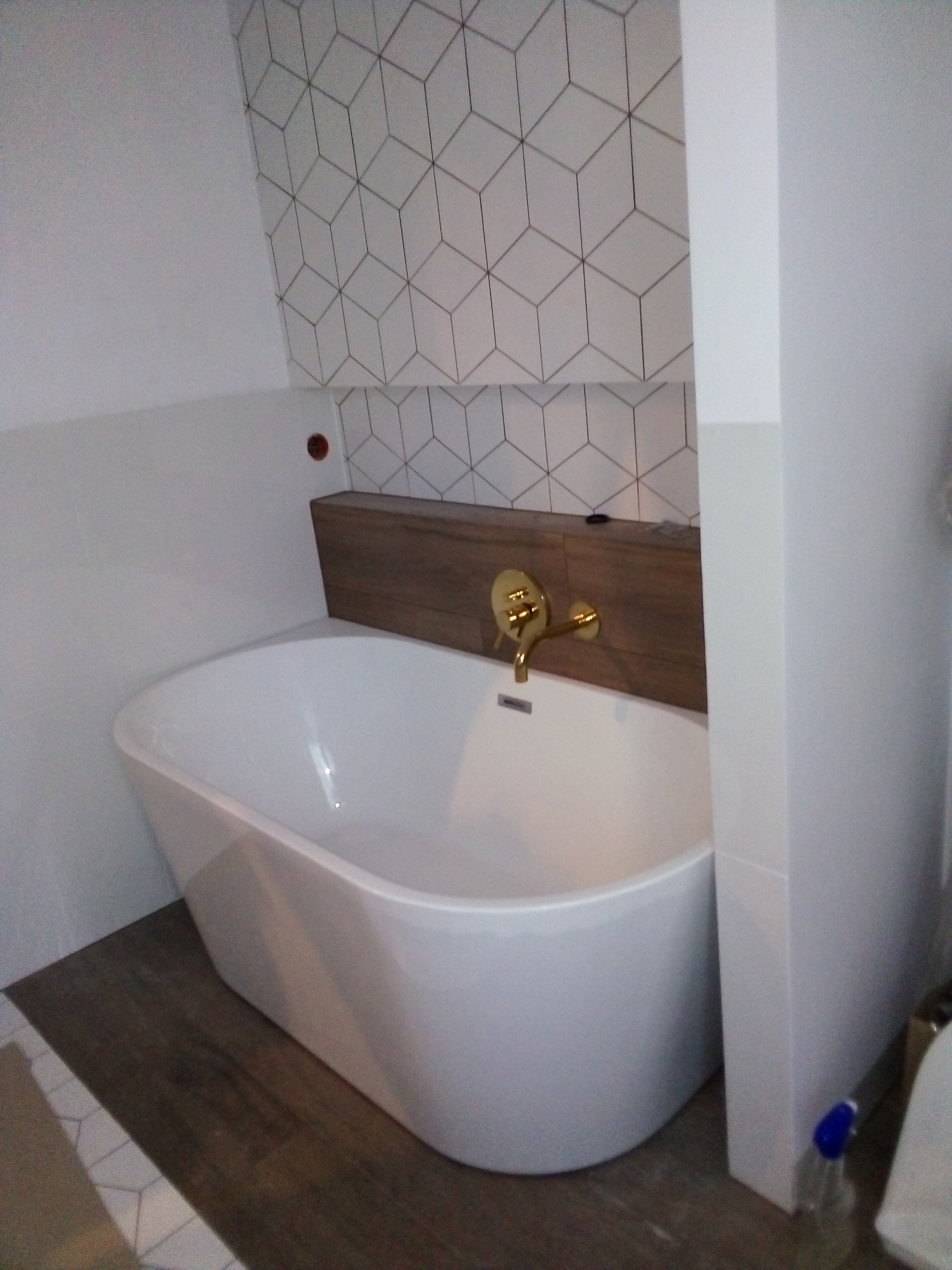 Nowoczesna łazienka z wolnostojącą białą wanną, złotą armaturą i geometrycznymi płytkami na ścianie, uzupełniona drewnianym panelem nad wanną.