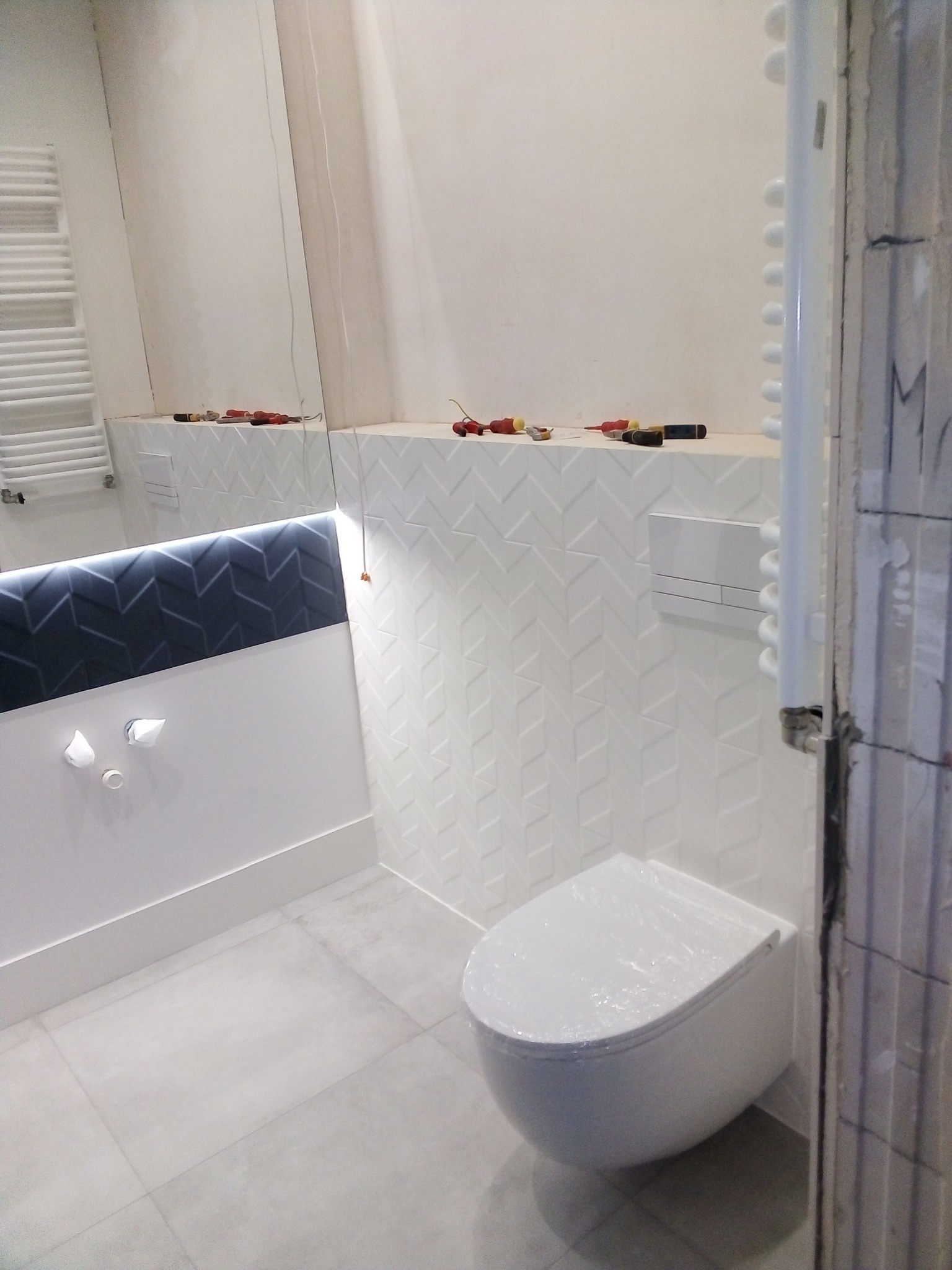 Nowoczesna łazienka w trakcie remontu, widoczne białe płytki ułożone we wzór jodełki, podwieszana toaleta zabezpieczona folią, oraz narzędzia na półce, odbijające się w lustrze z podświetleniem LED.