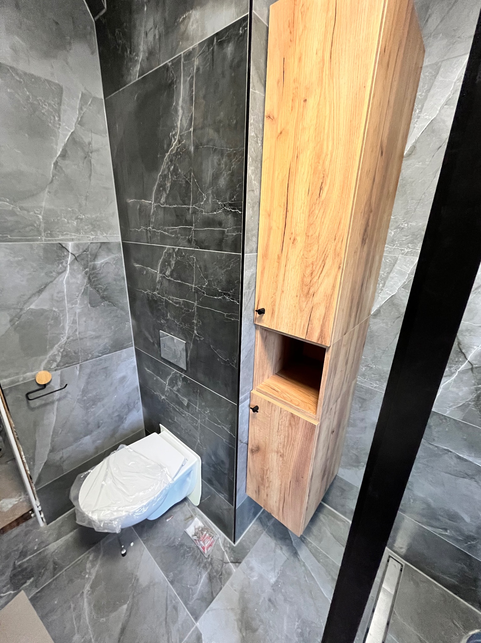 Wykończona łazienka z szarą marmurową okładziną, drewnianą szafką i toaletą zabezpieczoną folią. Widok z góry podkreśla nowoczesny design i dbałość o detale.