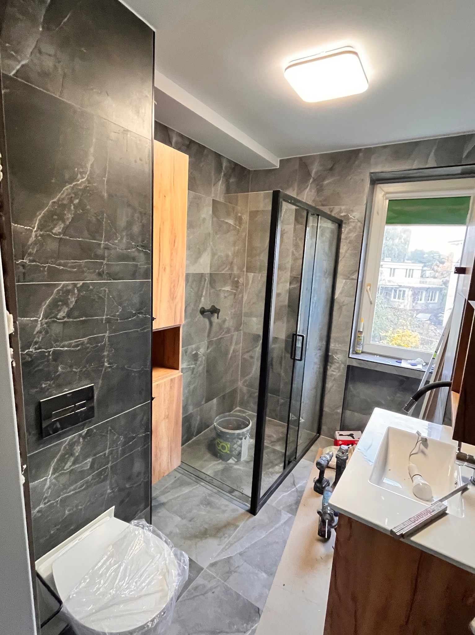 Nowoczesna łazienka w trakcie wykończenia: szare płytki imitujące marmur, kabina prysznicowa z czarnymi profilami, drewniana szafka i narzędzia. Widok na okno.