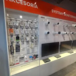 Ekspozycja w sklepie z systemami monitoringu: kamery różnych typów i rejestratory na białych półkach, widoczne logo Dahua, Kenik i Hikvision, oraz akcesoria w opakowaniach na ścianie po lewej.
