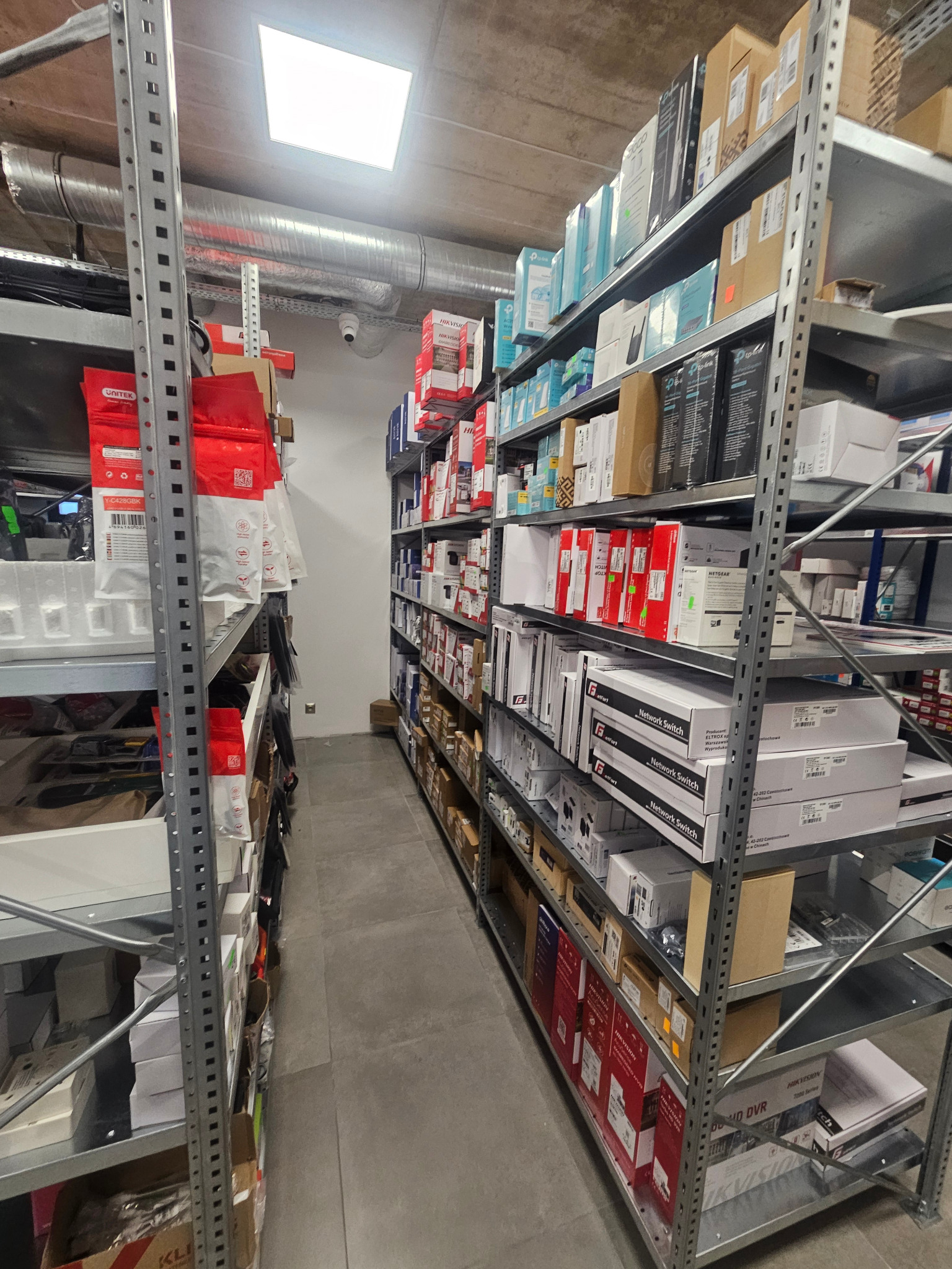 Magazyn z półkami regałowymi wypełnionymi pudełkami z urządzeniami elektronicznymi, w tym przełącznikami sieciowymi, rejestratorami DVR i akcesoriami, widoczne marki Netgear, Hikvision i Unitek.