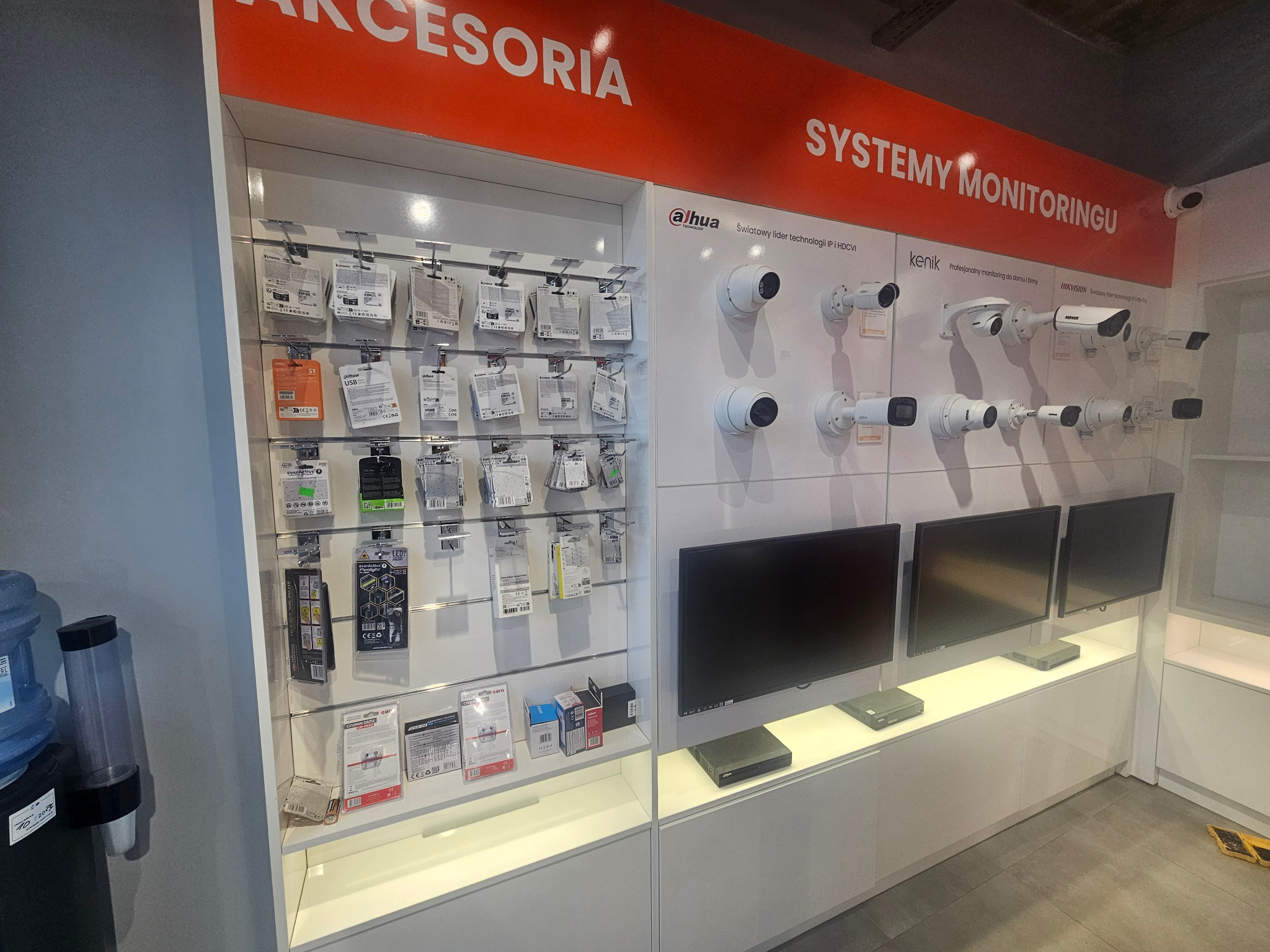 Ekspozycja w sklepie z systemami monitoringu: kamery różnych typów i rejestratory na białych półkach, widoczne logo Dahua, Kenik i Hikvision, oraz akcesoria w opakowaniach na ścianie po lewej.