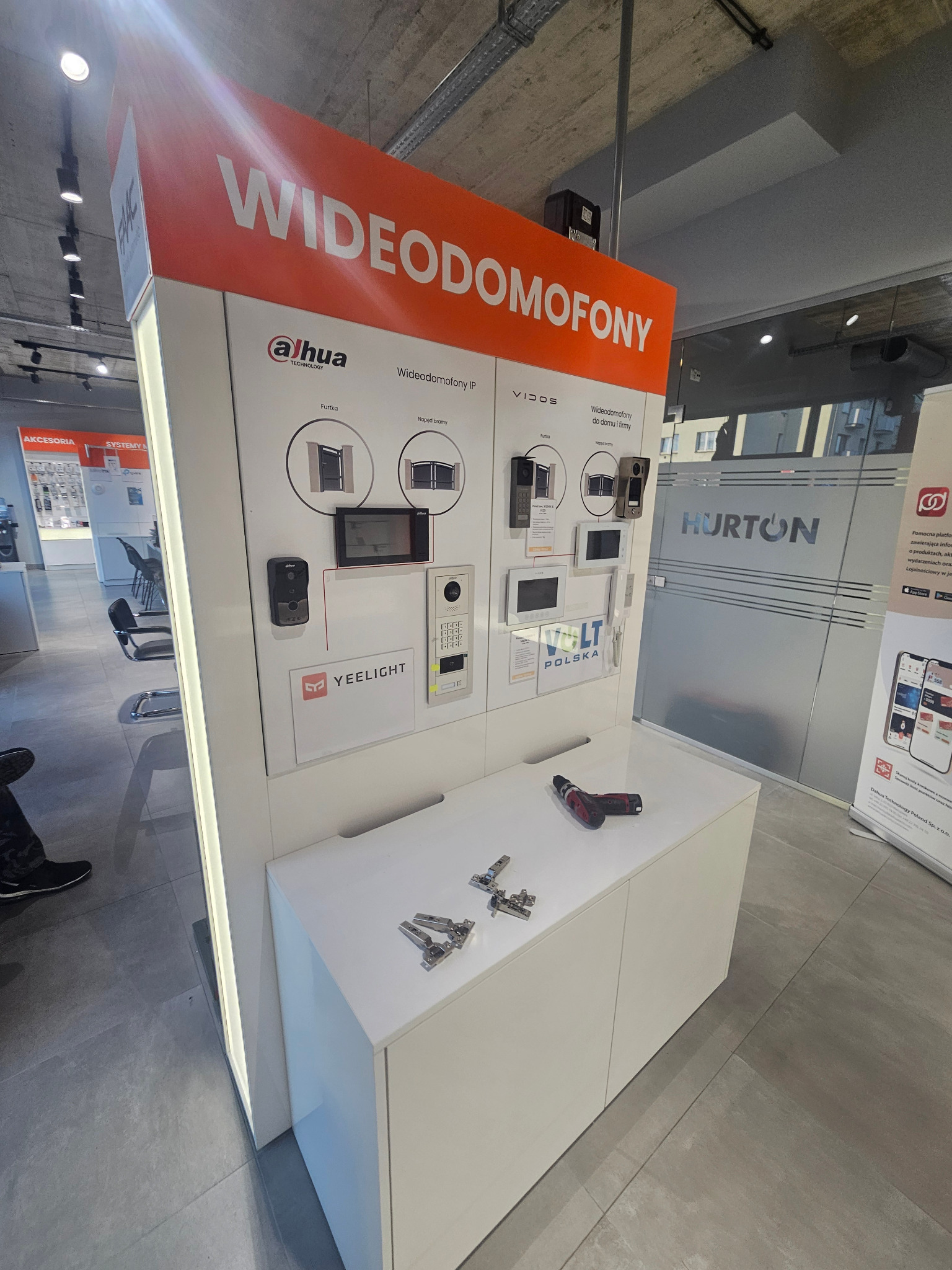 Ekspozycja różnych modeli wideodomofonów marek Dahua, Yeelight i Vidos w sklepie, z akcesoriami montażowymi i wkrętarką na blacie.