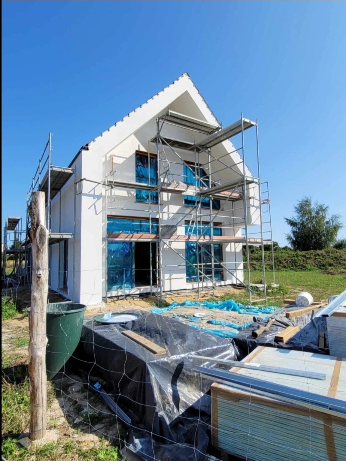 Nowoczesny dom w trakcie budowy, z białymi ścianami i rusztowaniami. Okna zabezpieczone folią, widoczne materiały budowlane i fragment zielonego otoczenia.