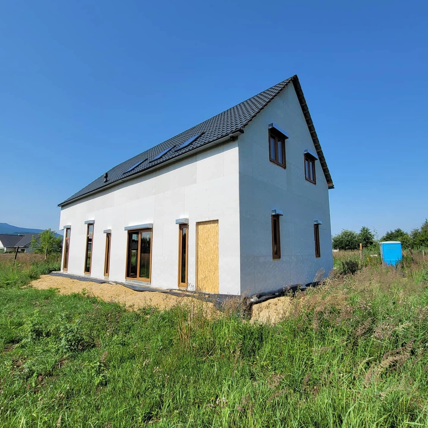 Surowy dom szkieletowy z brązowymi oknami i drzwiami zabezpieczonymi płytą, na tle nieba i zieleni, z widoczną górą w oddali. Etap budowy.