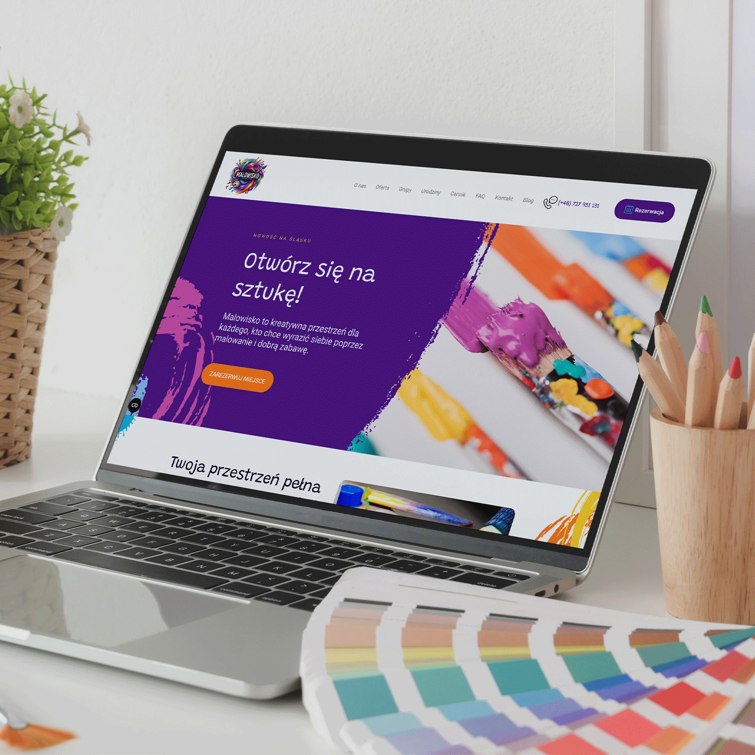 Laptop z otwartą stroną internetową Malowisko.art, prezentujący moduł rezerwacji. Obok paleta barw i kredki w drewnianym pojemniku na białym biurku.