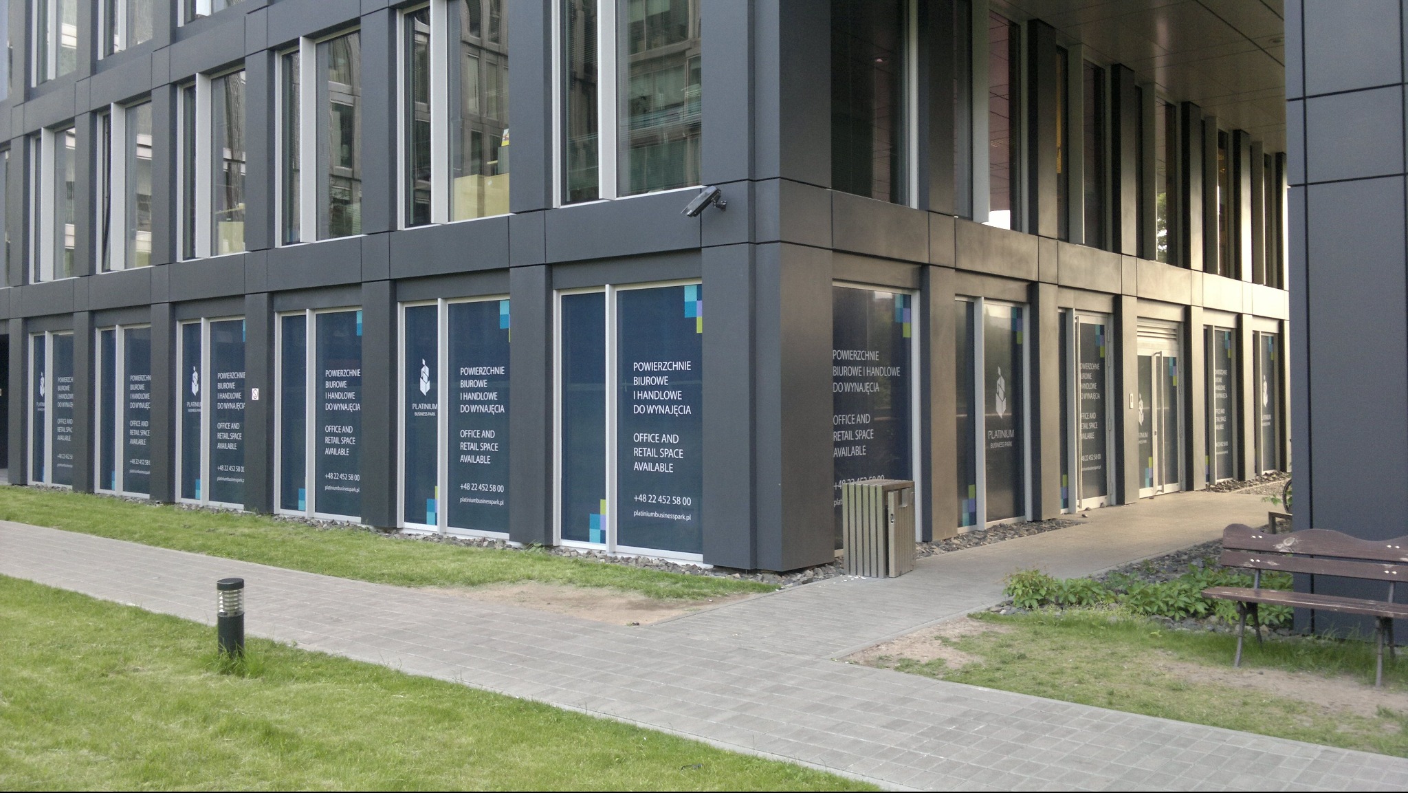 Zewnętrzny widok nowoczesnego biurowca z szarymi panelami i dużymi oknami, na których umieszczono informacyjne grafiki o dostępnych powierzchniach biurowych i handlowych na wynajem, z widocznym...