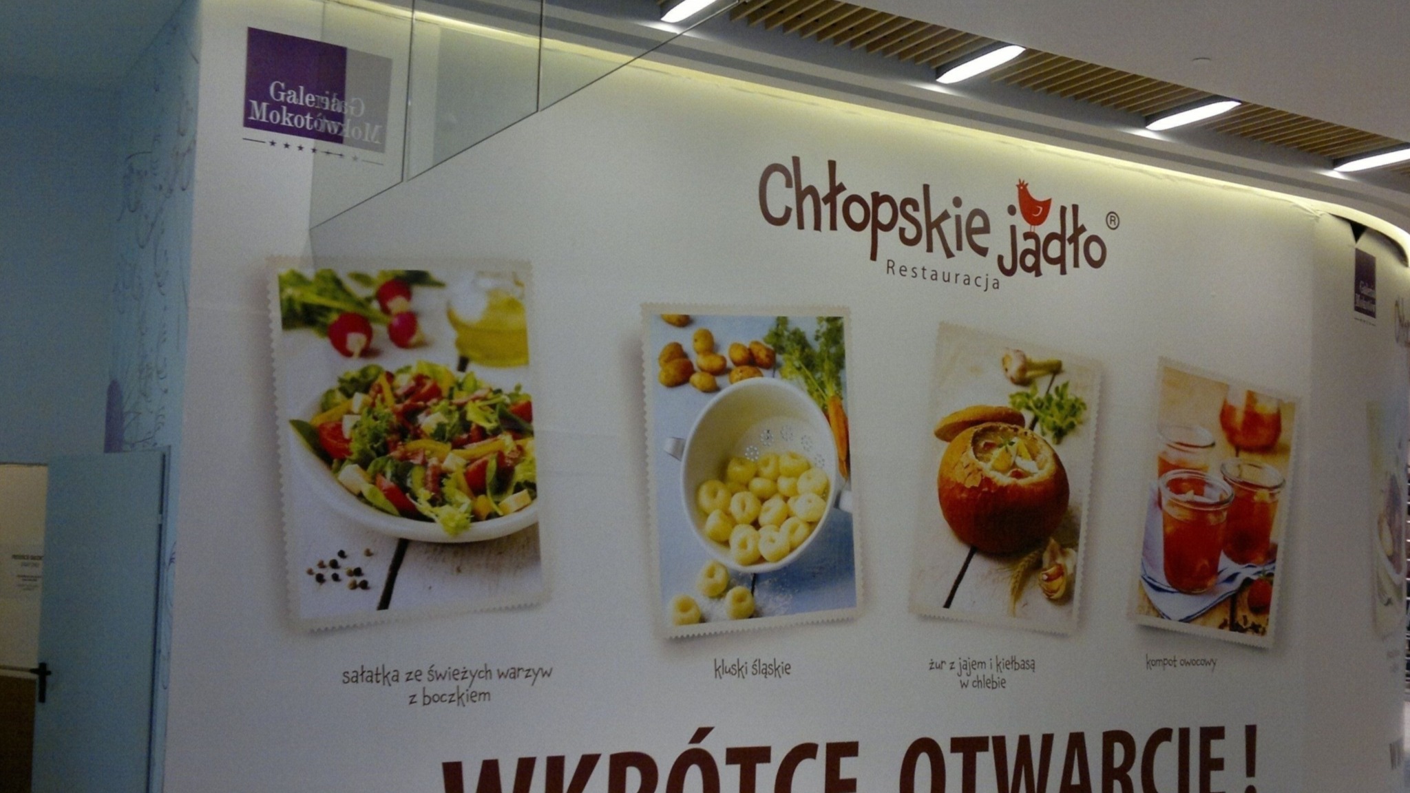 Wielkoformatowa naklejka reklamowa restauracji Chłopskie Jadło z wizualizacjami potraw: sałatki, klusek śląskich, żuru w chlebie i kompotu owocowego, informująca o wkrótce otwarciu w Galerii Mokotów.