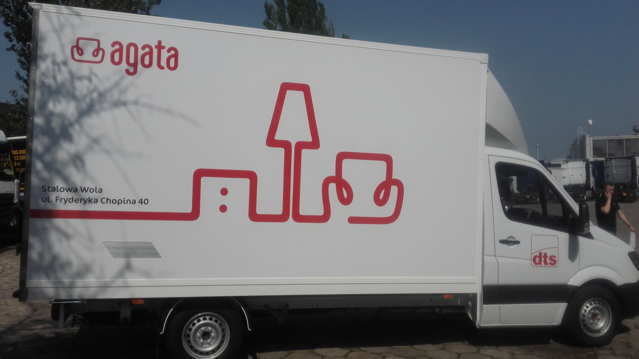 Biała furgonetka z czerwonym logo 'agata' i graficznym przedstawieniem mebli oraz adresem firmy Stalowa Wola, ul. Fryderyka Chopina 40, obok logo 'dts'.