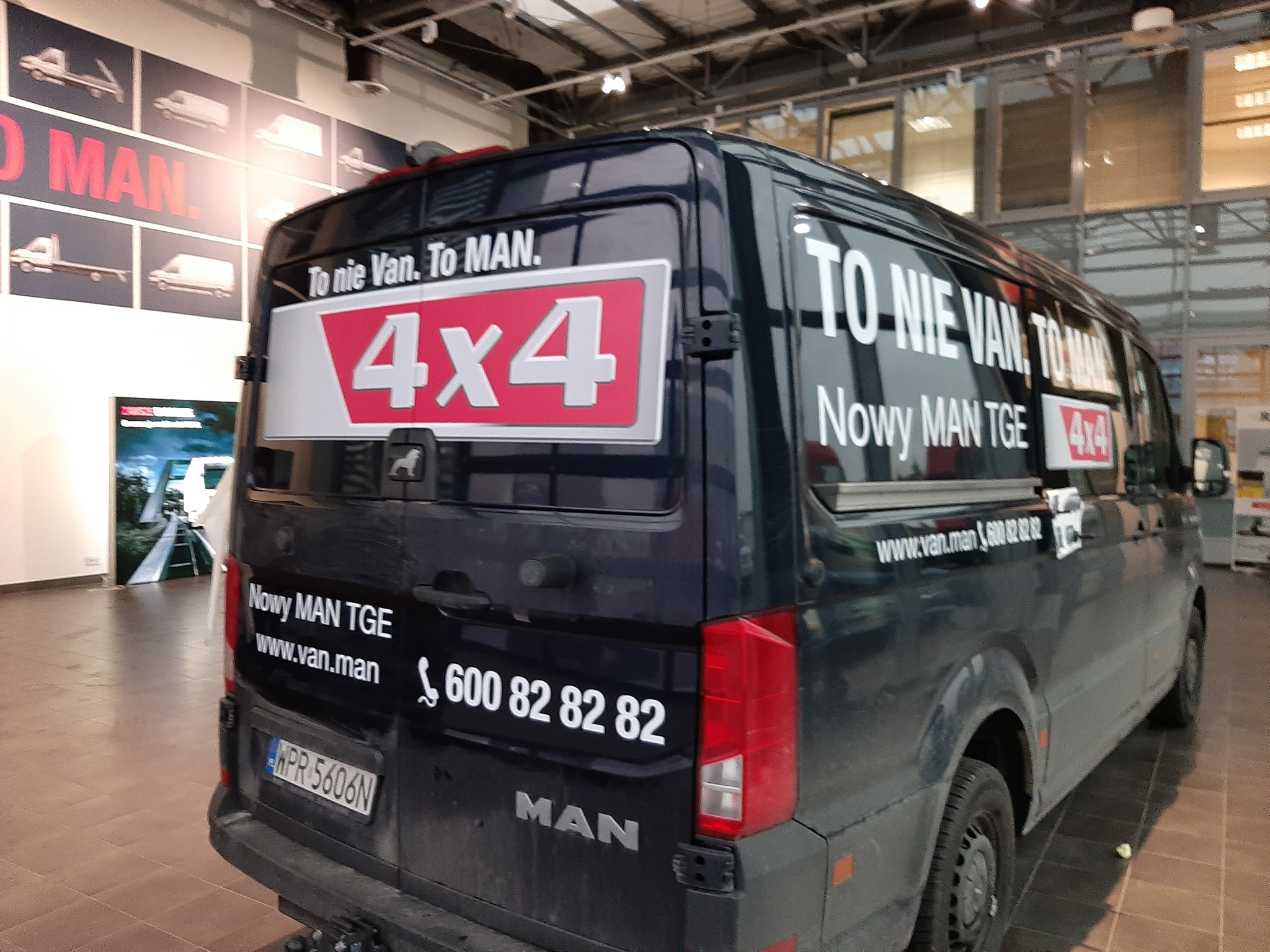Ciemny van MAN TGE z naklejkami reklamowymi 'To nie Van. To MAN.' i '4x4' na tylnej szybie i karoserii, prezentowany w salonie samochodowym.