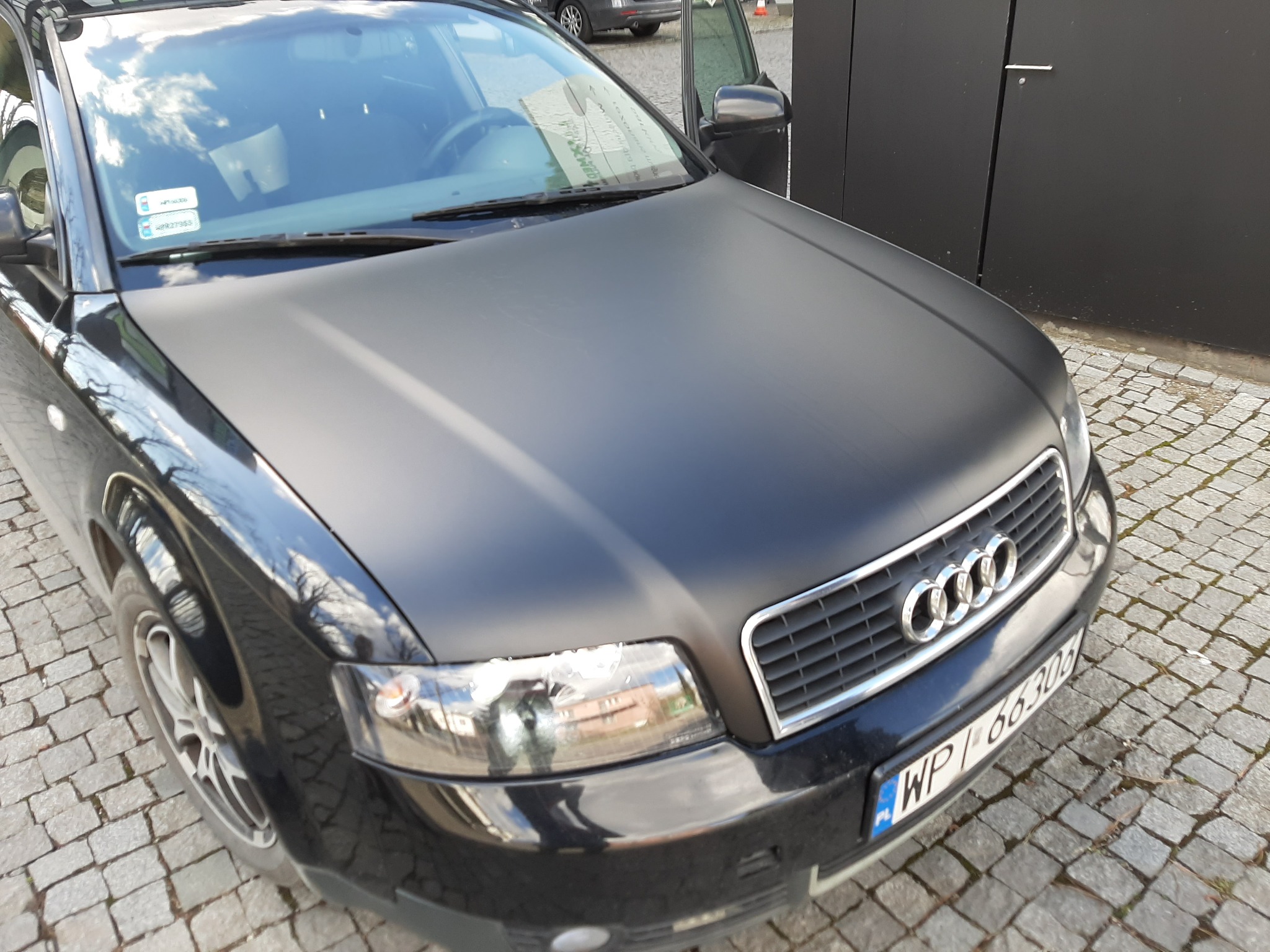 Czarny samochód Audi A4 z matową okleiną na masce, zaparkowany na kostce brukowej przed ciemną ścianą.