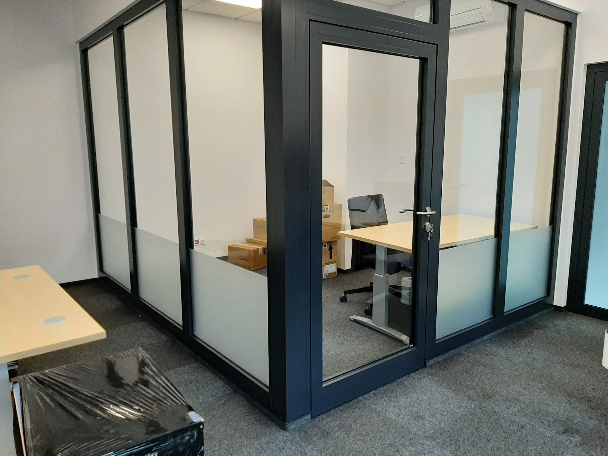 Nowoczesna szklana ścianka działowa z czarnymi ramami w biurze, dolna część szyb pokryta folią matującą, widoczne biurka i krzesło wewnątrz.