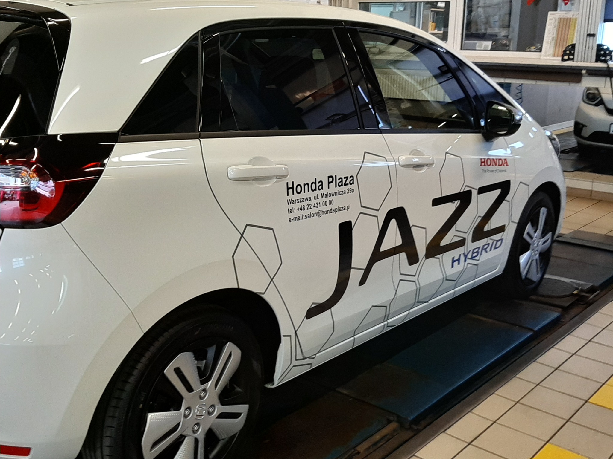Biała Honda Jazz Hybrid z czarnym napisem Jazz i logotypem Honda Plaza na boku, ozdobiona geometrycznym wzorem, stojąca na podnośniku w warsztacie.