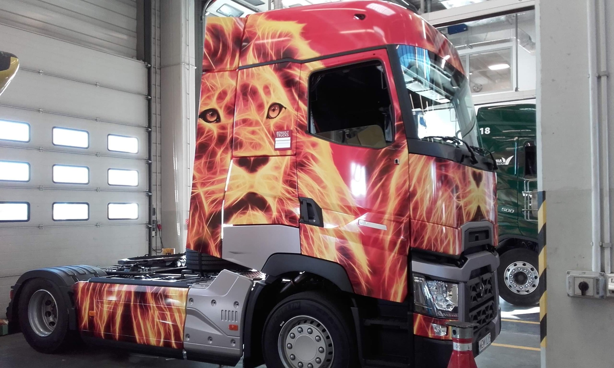 Czerwona ciężarówka z motywem płonącego lwa, zaparkowana w warsztacie w Piasecznie. Grafika na kabinie i boku pojazdu. W tle widoczna brama garażowa i fragment zielonej ciężarówki.