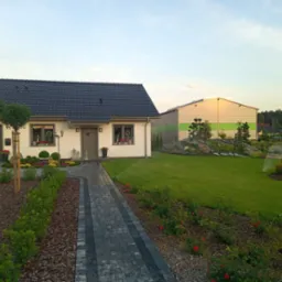 Nowoczesny dom z zadbanym ogrodem: trawnik z automatycznym nawadnianiem, ścieżka z kostki, rabaty z korą i ozdobnymi krzewami, formowane drzewa kuliste.