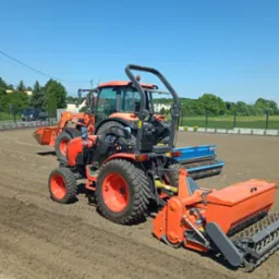 Pomarańczowy traktor Kubota z siewnikiem i przednim ładowaczem na przygotowanym, wyrównanym terenie pod zasiew trawy, błękitne niebo w tle.