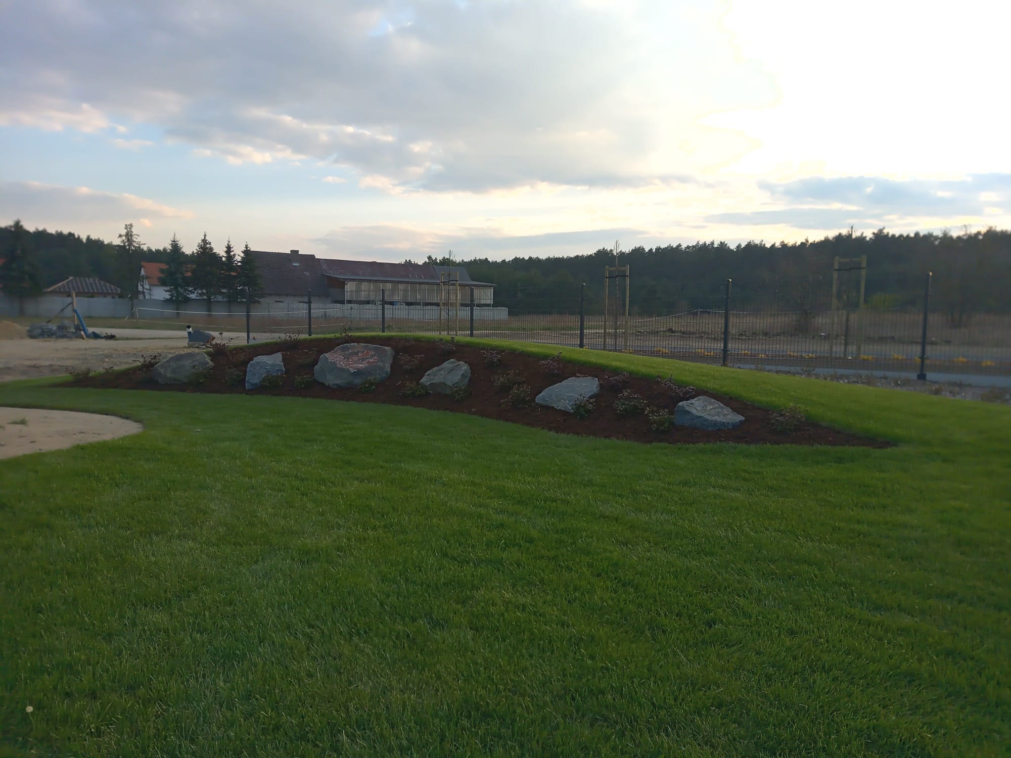 Nowo założony trawnik z rolki, z dekoracyjnym nasypem ziemnym obsadzonym małymi roślinami i dużymi kamieniami, w tle budynki gospodarcze i las.