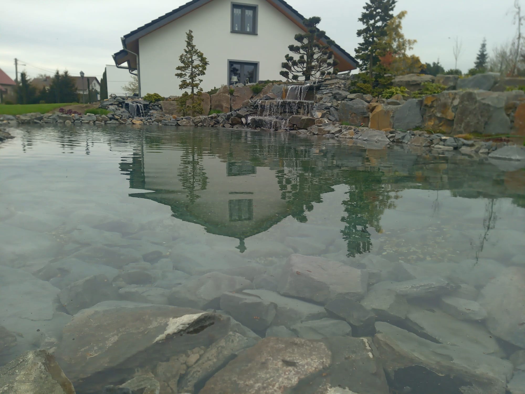 Oczko wodne z kaskadą wodną, otoczone kamieniami i roślinami, z odbiciem domu w wodzie.