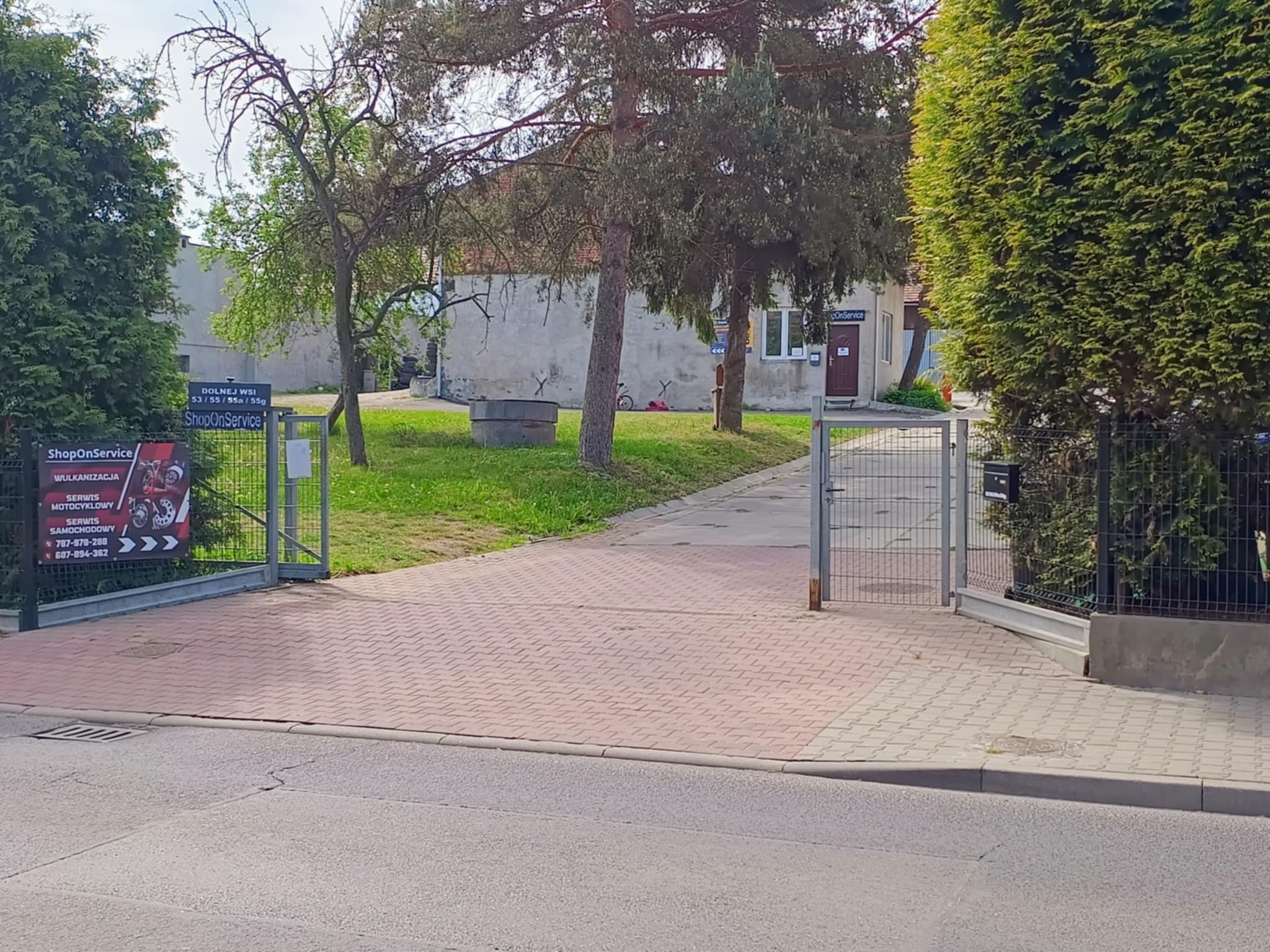 Widok na wjazd do warsztatu ShopOnService z reklamą wulkanizacji i serwisu motocyklowego, z budynkiem w tle. Otwarta brama wjazdowa.