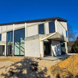 Surowy stan nowoczesnego domu jednorodzinnego z dużymi oknami i częściowo wykończoną elewacją z betonu komórkowego, widoczne rury spustowe i piaskowa ziemia wokół budynku.