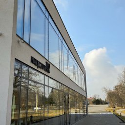 REVID SPÓŁKA Z OGRANICZONĄ ODPOWIEDZIALNOŚCIĄ - Nowoczesny budynek biurowy z dużą fasadą ze szkła refleksyjnego, na której widoczne jest odbicie drzew i nieba; na elewacji widnieje czarny napis '100 Nałęczowska'.