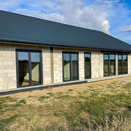 REVID SPÓŁKA Z OGRANICZONĄ ODPOWIEDZIALNOŚCIĄ - Surowy budynek w trakcie budowy z zamontowanymi grafitowymi oknami PCV, widok z zewnątrz na tle pola i błękitnego nieba z chmurami.