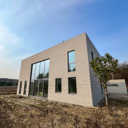 REVID SPÓŁKA Z OGRANICZONĄ ODPOWIEDZIALNOŚCIĄ - Nowoczesny dom z jasnej cegły z dużymi przeszkleniami, widok z poziomu gruntu na tle błękitnego nieba i suchych traw.
