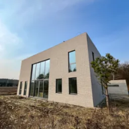 Nowoczesny dom z jasnej cegły z dużymi przeszkleniami, widok z poziomu gruntu na tle błękitnego nieba i suchych traw.