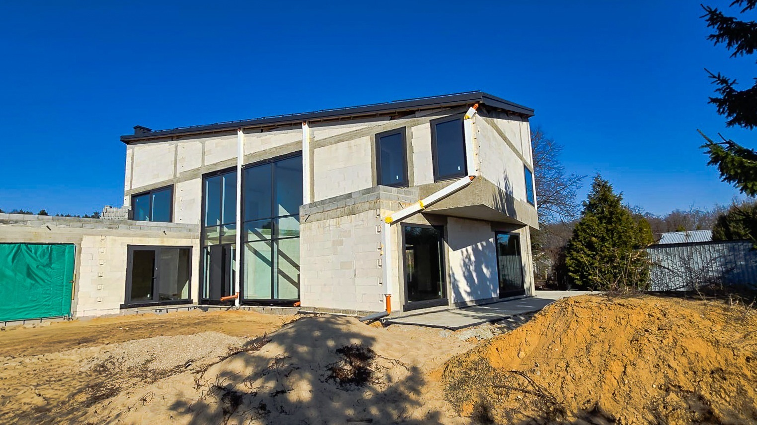 Surowy stan nowoczesnego domu jednorodzinnego z dużymi oknami i częściowo wykończoną elewacją z betonu komórkowego, widoczne rury spustowe i piaskowa ziemia wokół budynku.