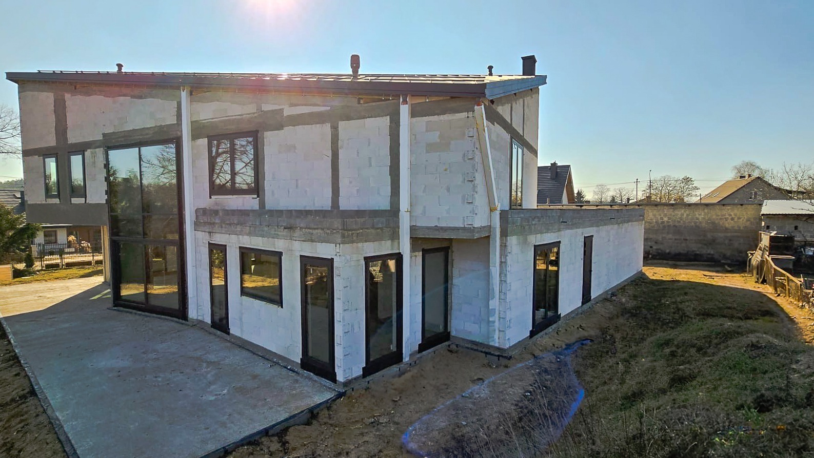 Nowoczesny dom w surowym stanie, z dużymi oknami z czarnymi ramami, widok z zewnątrz, teren wokół w trakcie prac ziemnych.