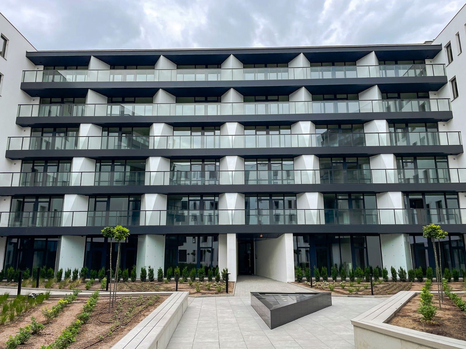 Nowoczesny budynek apartamentowy z balkonami z szklanymi balustradami i białymi elementami fasady, widok z poziomu gruntu na zagospodarowany teren z niską zielenią.