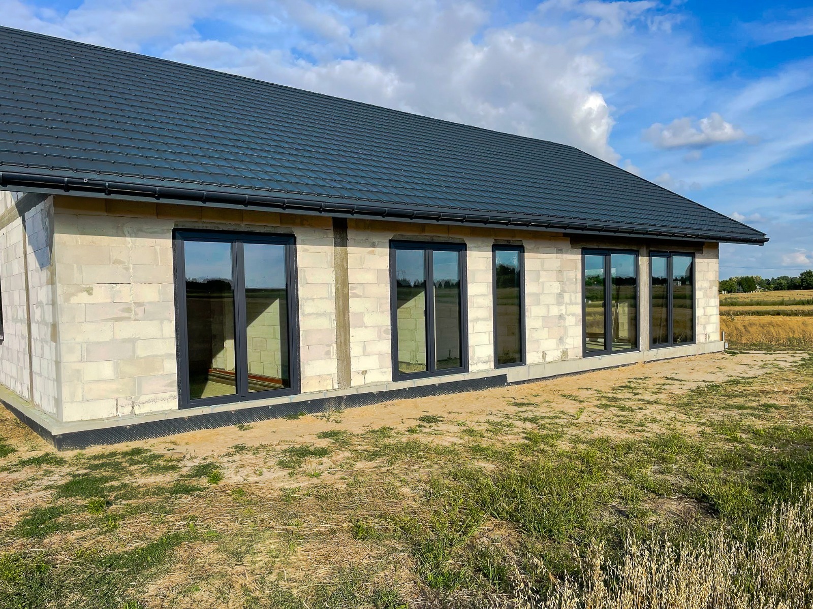 Surowy budynek w trakcie budowy z zamontowanymi grafitowymi oknami PCV, widok z zewnątrz na tle pola i błękitnego nieba z chmurami.
