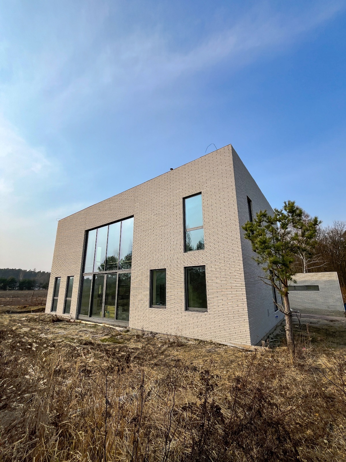 Nowoczesny dom z jasnej cegły z dużymi przeszkleniami, widok z poziomu gruntu na tle błękitnego nieba i suchych traw.