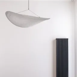 Minimalistyczne wnętrze z wiszącą lampą w kształcie liścia i czarnym grzejnikiem żeberkowym na tle białej ściany i drewnianej podłogi.