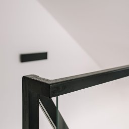 FILIP RAUPUK ARCHITEKTURA - Minimalistyczna czarna balustrada szklana na tle białej ściany z czarnym elementem dekoracyjnym. Ujęcie z bliska, akcentujące detale wykończenia drewna.