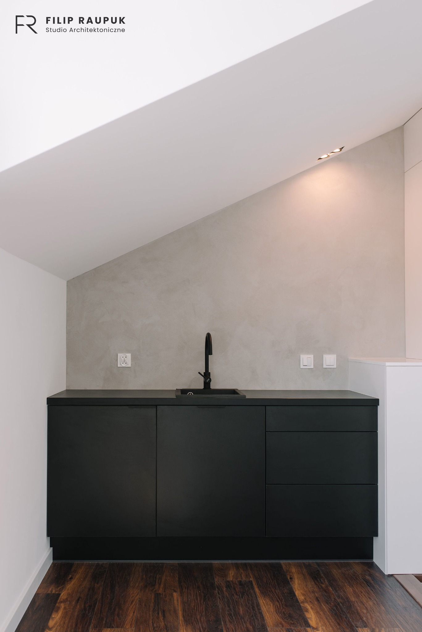 Czarne szafki kuchenne z matowym wykończeniem pod skośnym sufitem, minimalistyczna armatura, drewniana podłoga. Widoczne gniazdka i włączniki na ścianie.