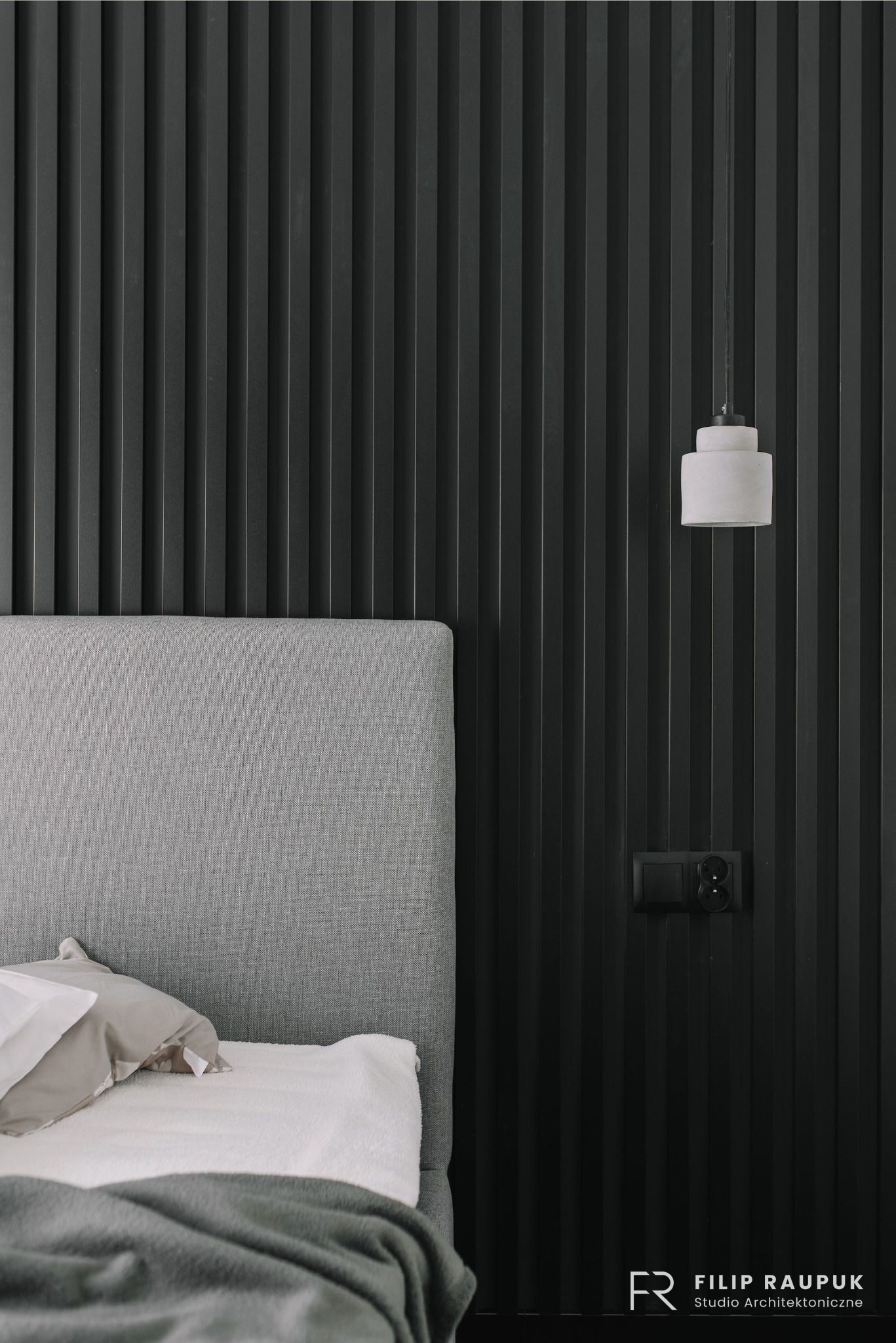 Minimalistyczna sypialnia w Szczecinie: łóżko z szarym zagłówkiem na tle ściany z czarnych paneli, lampka nocna i gniazdko. Subtelne oświetlenie.