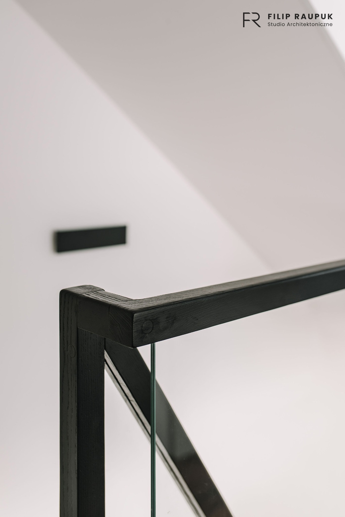 Minimalistyczna czarna balustrada szklana na tle białej ściany z czarnym elementem dekoracyjnym. Ujęcie z bliska, akcentujące detale wykończenia drewna.