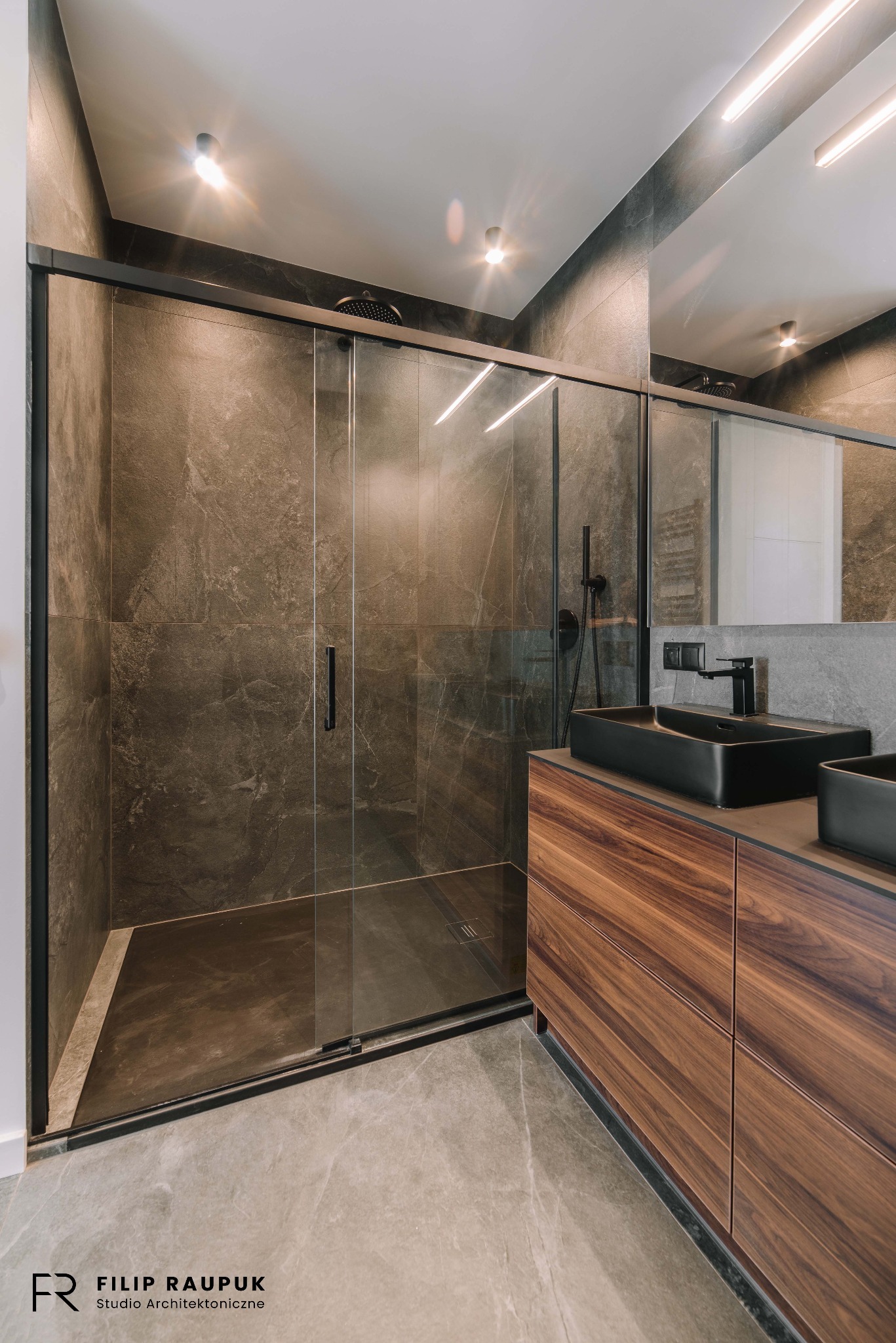 Nowoczesna łazienka z kabiną typu walk-in, ciemnymi płytkami imitującymi kamień i drewnianą szafką pod umywalką. Minimalistyczny design z czarnymi akcentami.