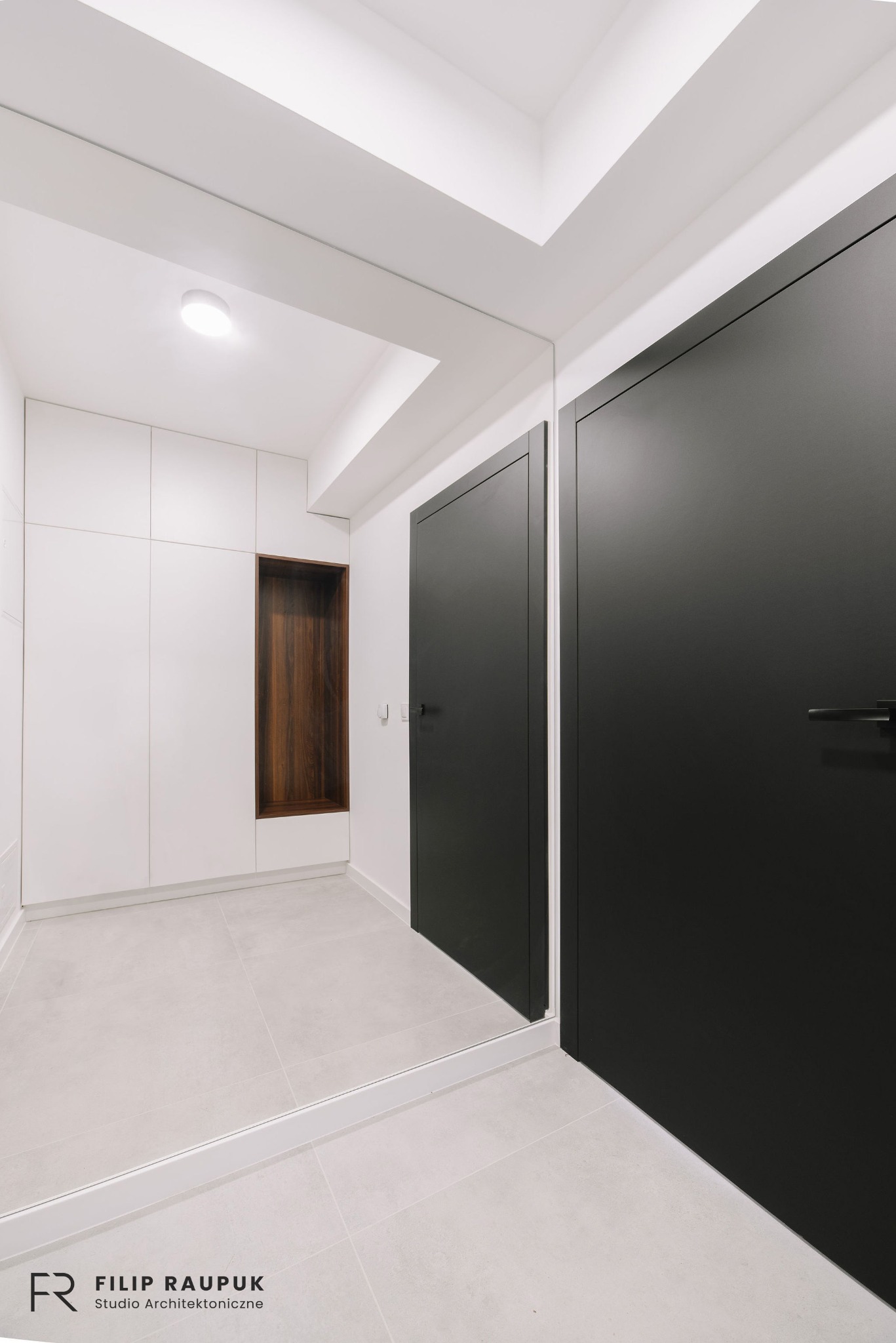 Wnętrze: Minimalistyczny przedpokój z szafą w zabudowie, drewnianą wnęką i dużym lustrem. Czarne drzwi dodają kontrastu, a jasna podłoga i ściany powiększają przestrzeń.