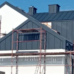 Montaż grafitowej blachy na dachu domu jednorodzinnego, widoczne rusztowania i kominy wentylacyjne.