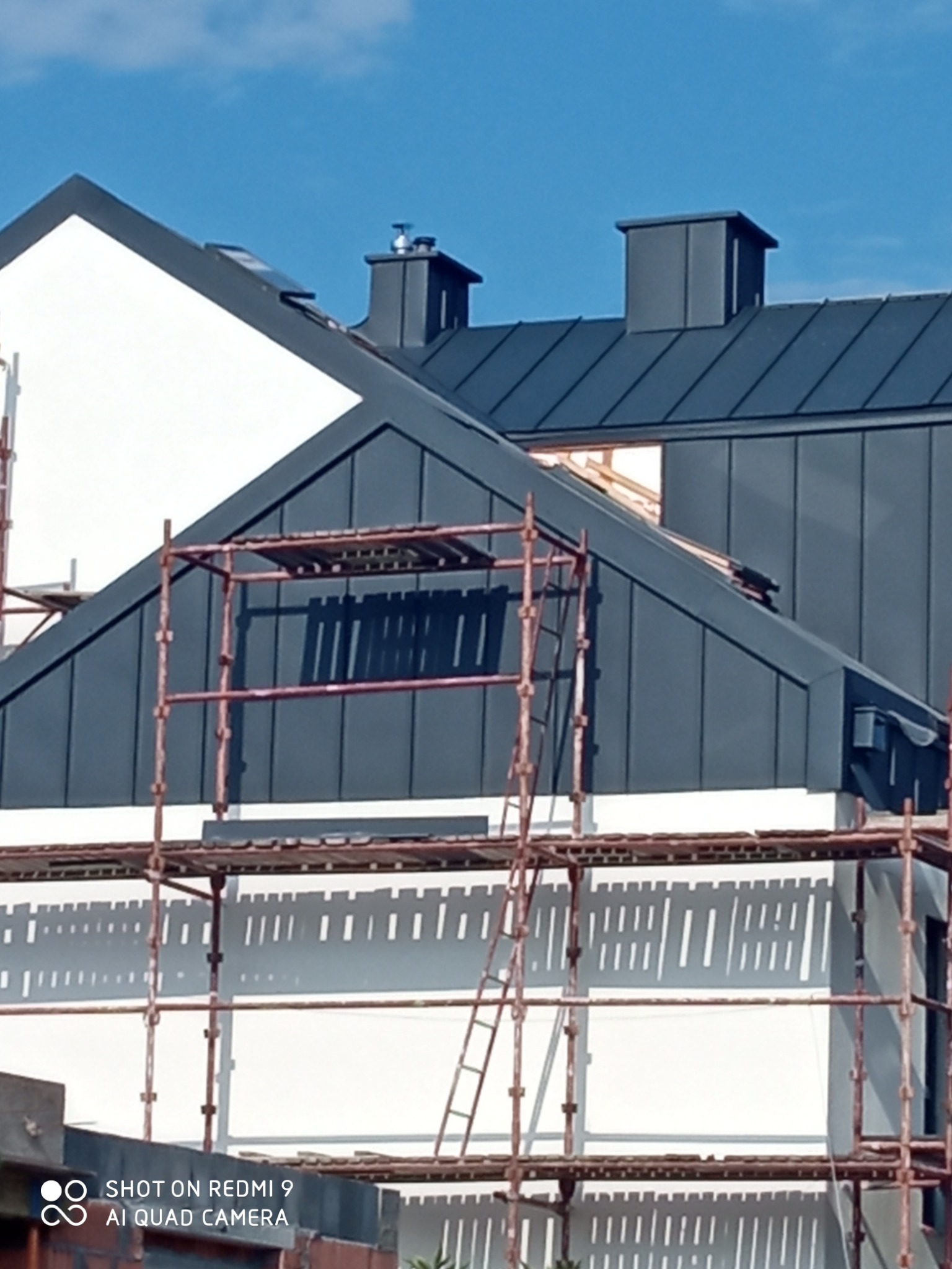 Montaż grafitowej blachy na dachu domu jednorodzinnego, widoczne rusztowania i kominy wentylacyjne.