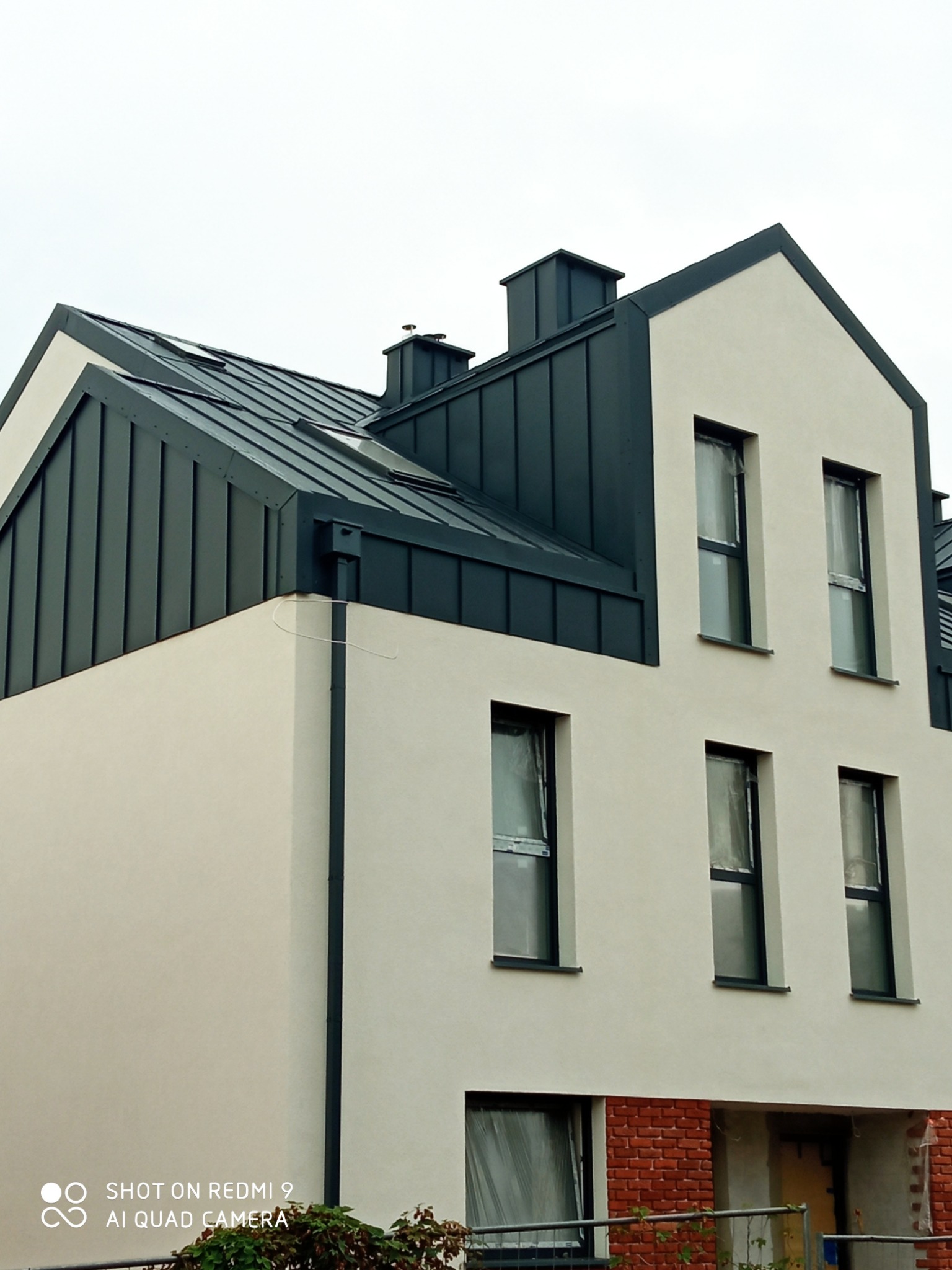 Nowoczesny dom jednorodzinny z ciemnoszarą blachą na dachu i jasną elewacją, okna zabezpieczone folią ochronną, widoczny komin i fragment muru z czerwonej cegły w dolnej partii budynku.